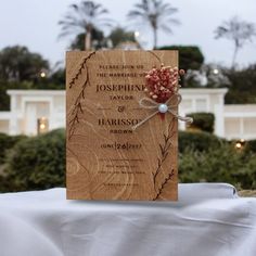 a wooden wedding card on top of a white table cloth with flowers in the middle