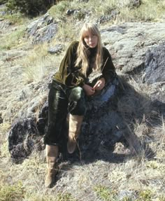 a woman sitting on top of a large rock in the middle of a grass covered field