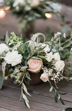 an arrangement of white flowers and greenery on a wooden table with candles in the background