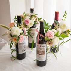 three wine bottles with flowers and greenery in them sitting on a white table cloth