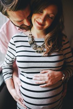 a pregnant couple cuddles in front of a window