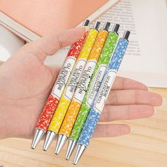 four different colored pens in the palm of a person's hand next to an open book