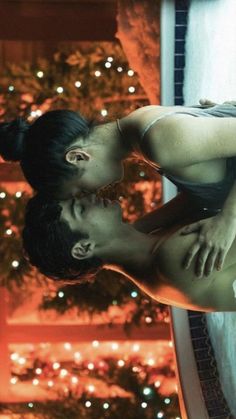 a man and woman are kissing in front of a christmas tree with lights on it