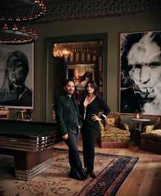 a man and woman standing next to each other in a living room with pictures on the wall