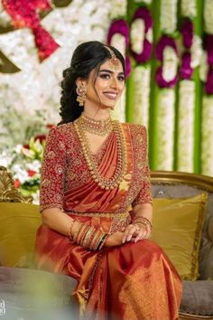 a woman in an orange and gold sari sitting on a couch with flowers behind her