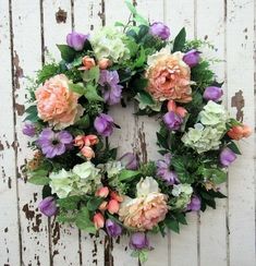 a wreath with pink and purple flowers hanging on a white wooden wall, surrounded by greenery