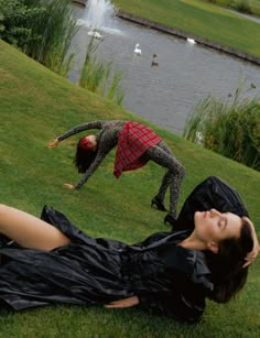 two women laying on the grass in front of a pond