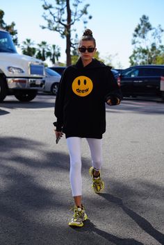 a woman walking down the street wearing yellow shoes and a black sweatshirt with smiley face on it