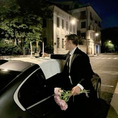 a man in a tuxedo sitting on the back of a car holding flowers