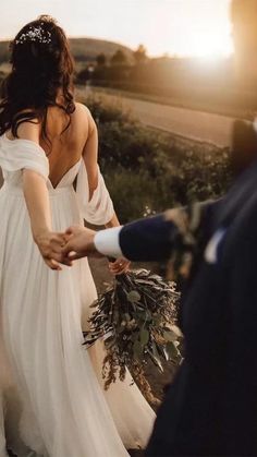 a bride and groom holding hands walking down a dirt road in front of the sun