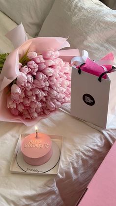 a pink birthday cake sitting on top of a bed next to a bouquet of flowers