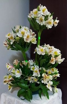 white flowers are in a vase on a small tableclothed stand with a wall behind it