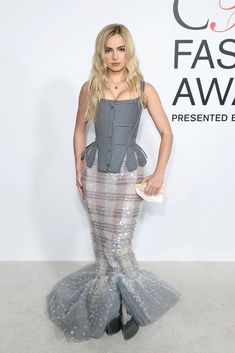 a woman in a gray and silver dress standing on a white carpet with her hands behind her back