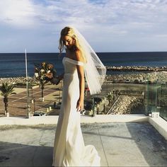 a woman in a wedding dress standing on a balcony looking at the ocean with her veil blowing in the wind