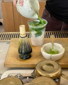 a person pouring green liquid into a cup on top of a wooden tray next to other items