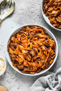 two bowls filled with pasta and meat on top of a table next to spoons