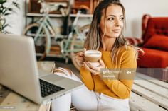 a woman sitting at a table holding a cup of coffee in front of her laptop