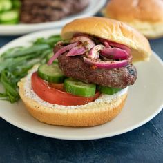 a hamburger with onions, lettuce and tomato on a white plate next to another burger