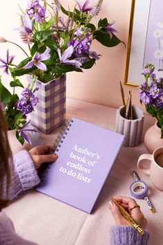 a woman sitting at a table writing on a notebook with purple flowers in the background