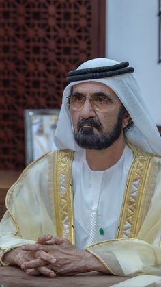 a man sitting at a table with his hands folded in front of him, wearing a white and gold outfit