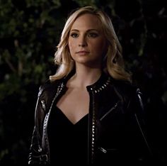 a woman in a black dress and leather jacket looking off into the distance with trees behind her