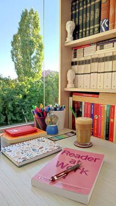 there is a book, cup and pen on the desk in front of the window