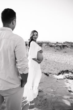 a pregnant woman standing on the beach with her husband looking at her in black and white