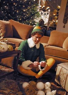 a man sitting on the floor in front of a christmas tree wearing a gnome hat