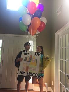 two people holding up a sign that says are you for hoco? with balloons in the background