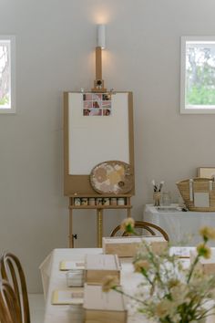 an easel is next to a white table with flowers on it in front of two windows