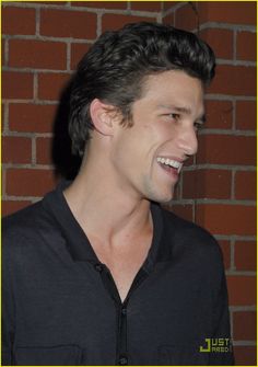 a smiling man in front of a brick wall with his hair pulled back and wearing a black shirt