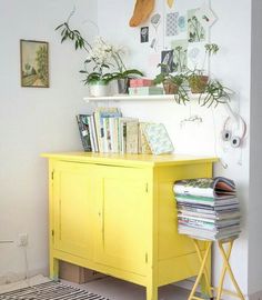 a yellow cabinet with books and plants on top in a room that has white walls