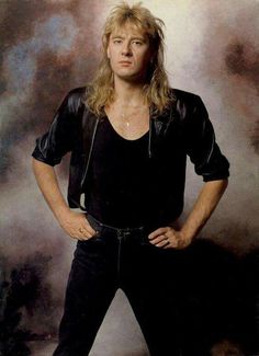 a man with long hair and black shirt posing for a photo in front of clouds