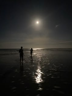 two people are walking on the beach at night with the moon in the sky above them