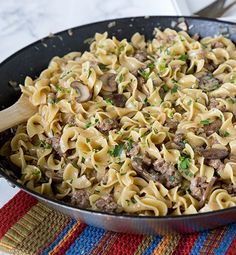 a skillet filled with pasta and meat on top of a colorful place mat next to silverware