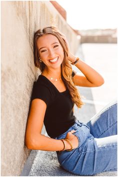 a beautiful young woman sitting on the side of a building smiling at the camera with her hands behind her head