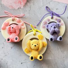 three crocheted teddy bears are hanging from small ornaments on a gray surface next to pink flowers