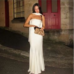 a woman in a white dress is holding a purse and standing on the street at night