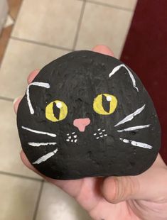 a hand holding a rock with a black cat painted on it's face and yellow eyes