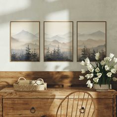 a wooden dresser topped with a vase filled with white flowers next to two framed pictures