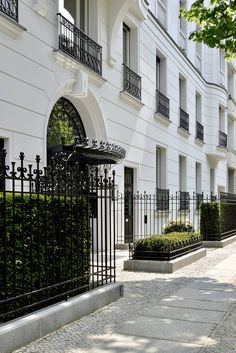 a white building with black iron fence and bushes on the sidewalk in front of it