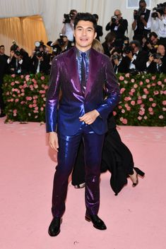 a man in a purple suit and tie standing on a pink carpet with people taking pictures behind him