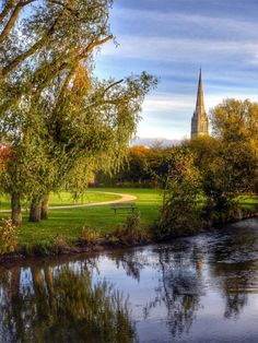 a river running through a lush green park next to a tall church tower in the distance