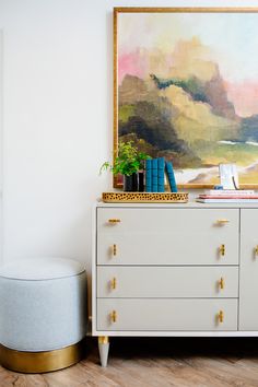 a white dresser with books and plants on it in front of a large art piece