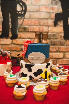 a cake and cupcakes on a red tablecloth with cow print, cowboy boots and other decorations