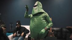 a man in a green dinosaur costume standing next to two men sitting on couches