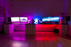 three computer monitors sitting on top of a wooden desk in front of two purple and red lights