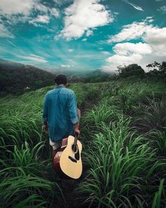 a man walking through tall grass with a guitar in his hand and the sky above him