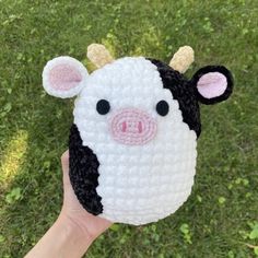 a hand holding up a small crocheted cow toy in the middle of grass