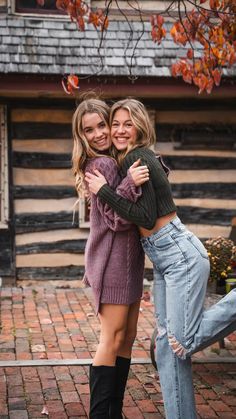 two young women hugging each other in front of a log cabin with autumn leaves on the ground